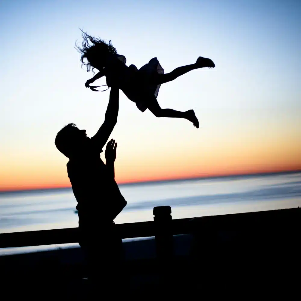 Silhouette d’un père lançant son enfant dans les airs au coucher du soleil : la place du père, la transmission et la crise du masculin